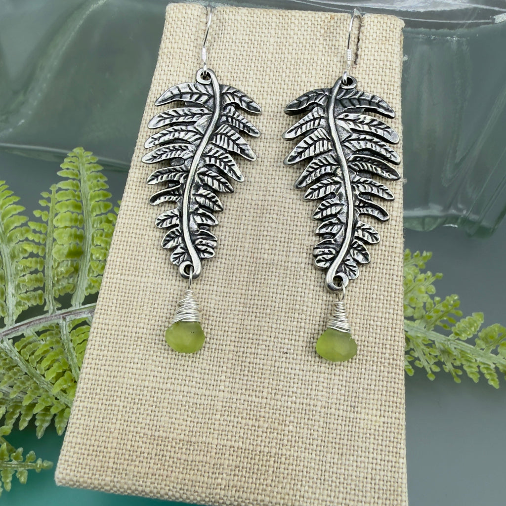 Silver fern frond or leaf shaped earrings with green peridot drops on a beige display with fern leaves in the background.