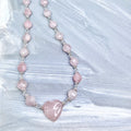 Necklace with a 3/4" pink heart-shaped center bead, with smaller faceted flat round beads with silver and clear iridescent crystal accents on a textured light grey background