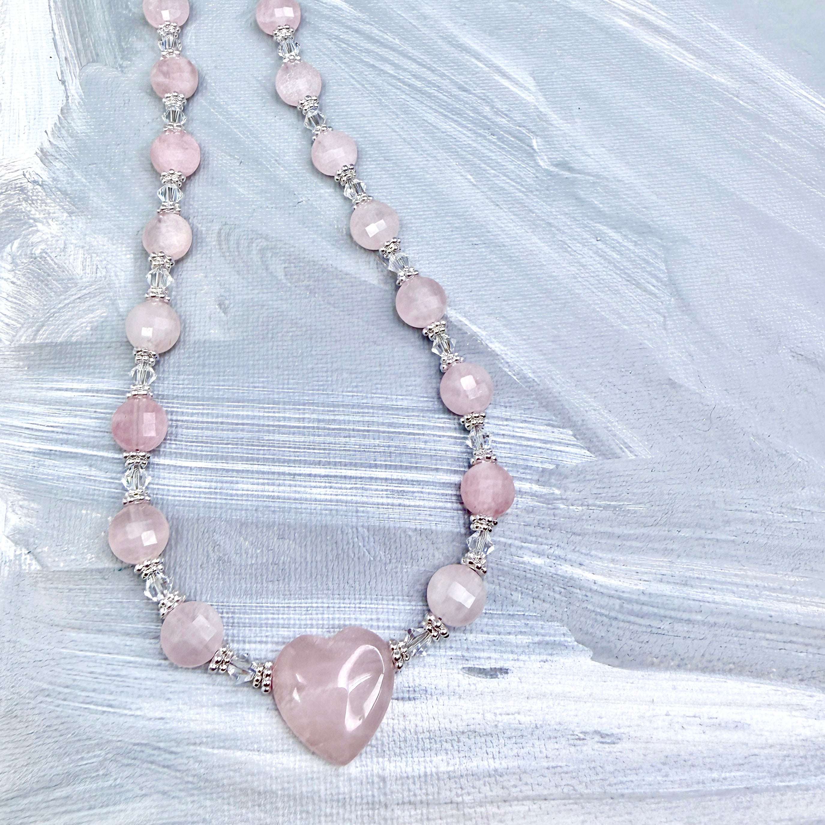 Necklace with a 3/4" pink heart-shaped center bead, with smaller faceted flat round beads with silver and clear iridescent crystal accents on a textured light grey background