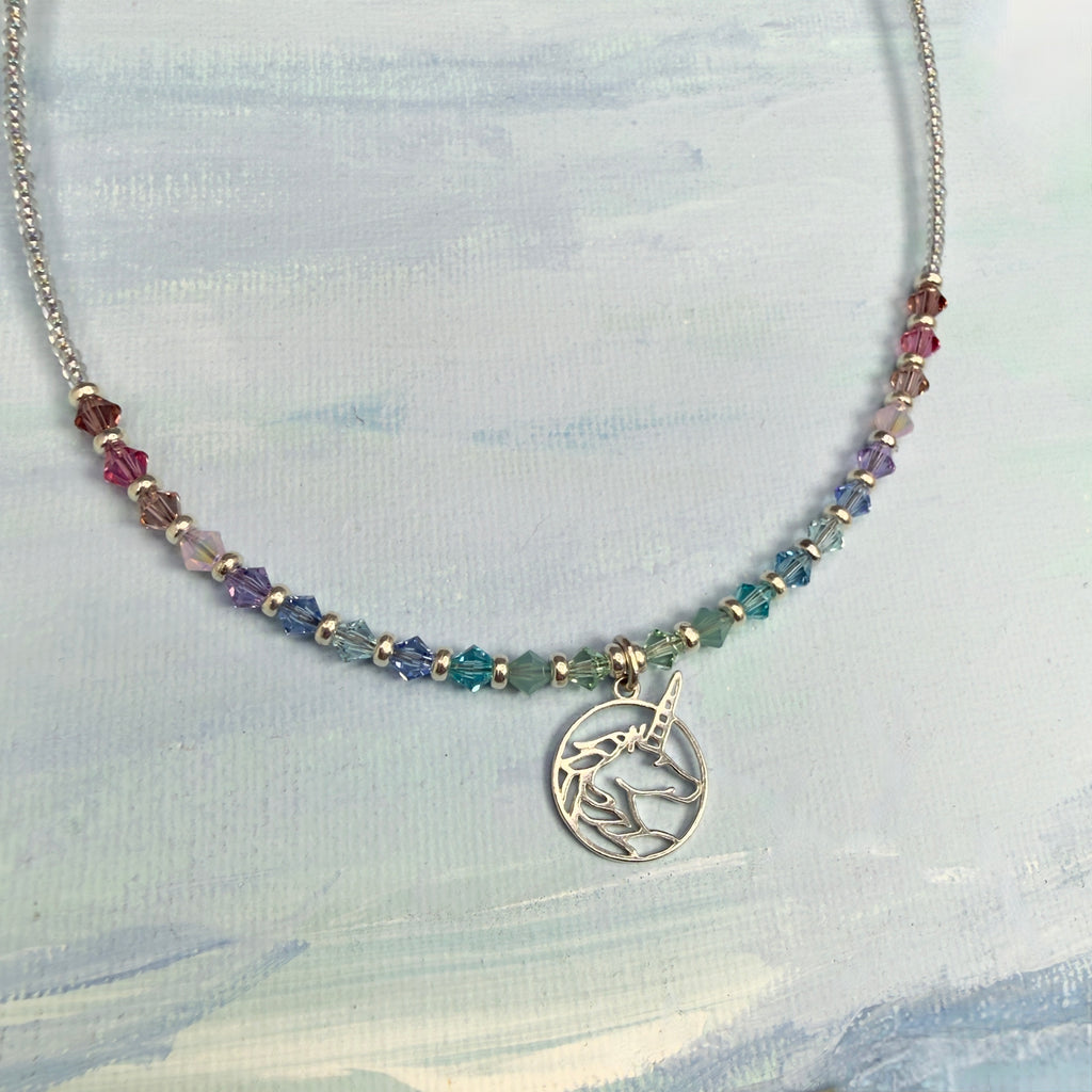 Necklace with multicolored pastel crystal beads and a circular silver unicorn pendant on a light blue textured background