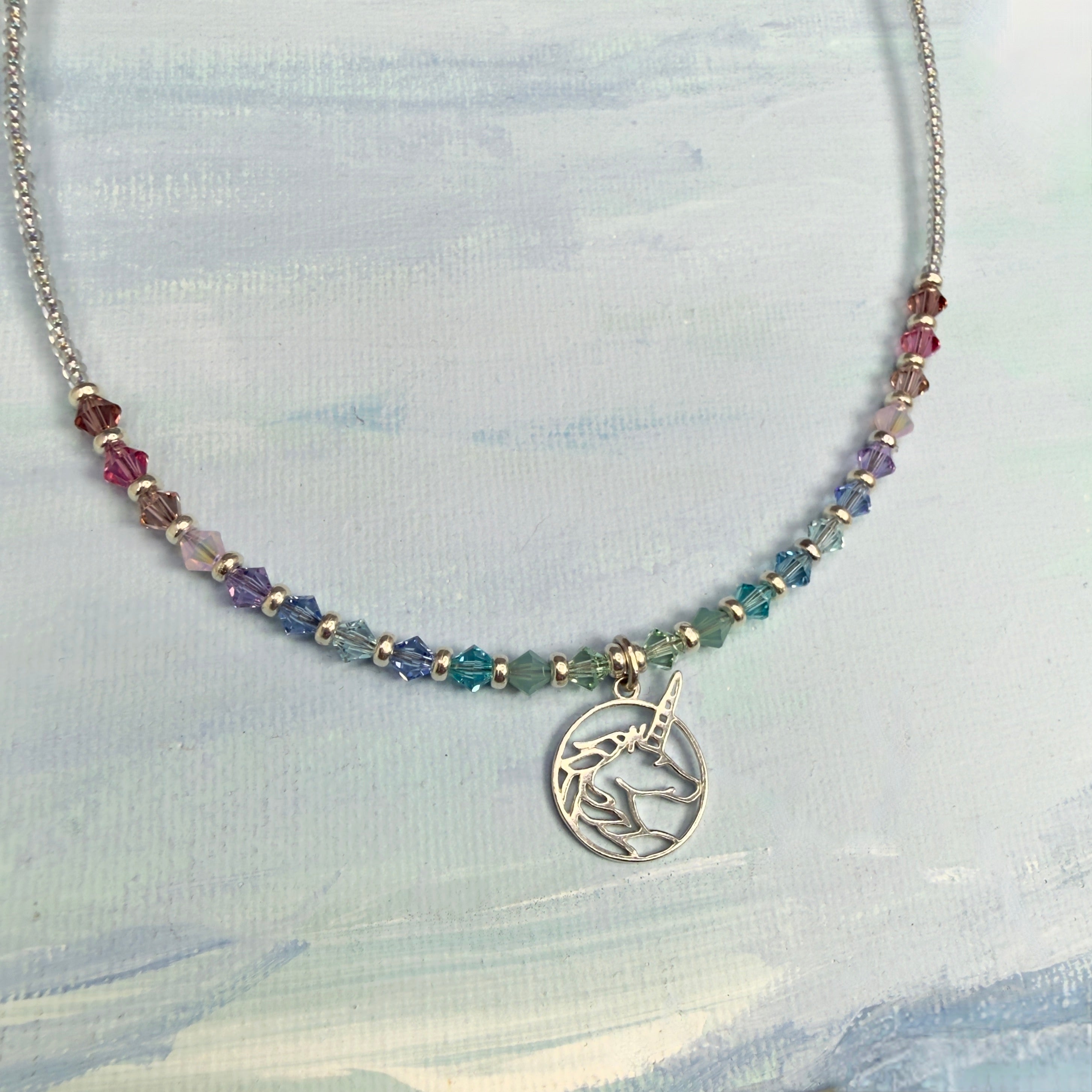 Necklace with multicolored pastel crystal beads and a circular silver unicorn pendant on a light blue textured background