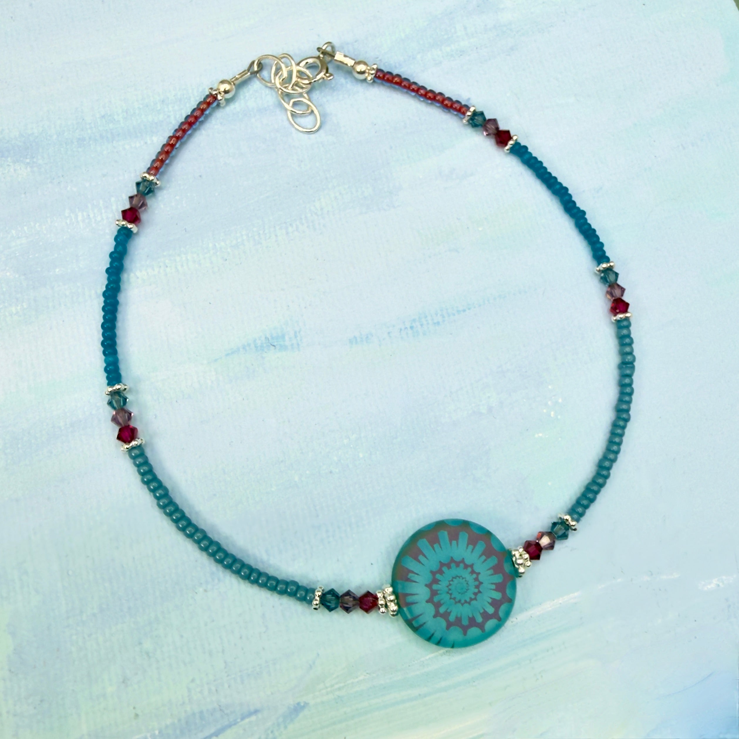 Beaded anklet  in cerulean blue and fuchsia with a center spiral disc bead on a light blue textured background