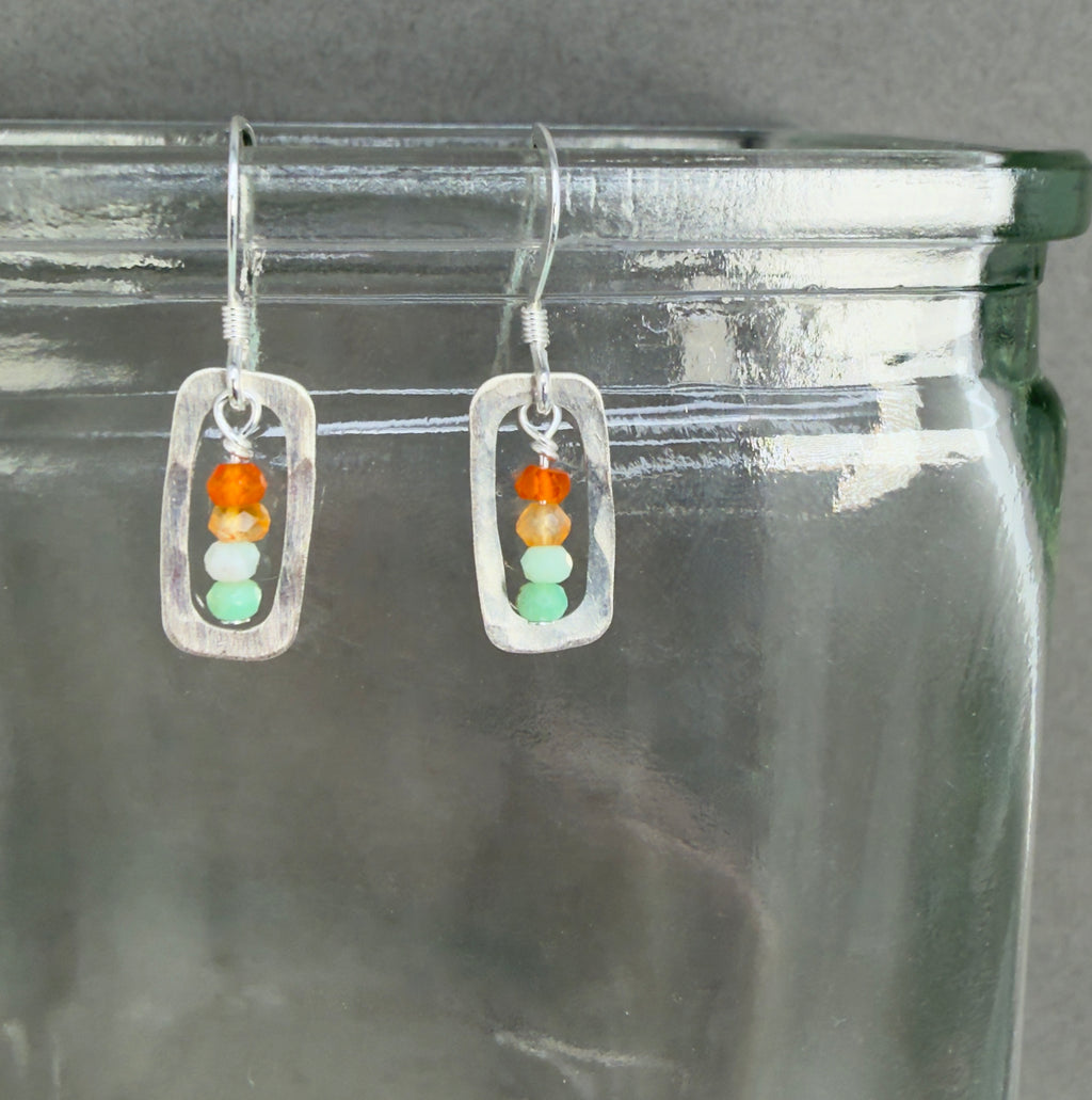 Pair of earrings with a stack or orange, pale orange, pale minty green and medium minty green beads framed with a rounded silver rectangular frame, on sterling silver hooks. They're shown hanging on a clear glass jar with that hint of green clear glass has, in front of a dark grey background.