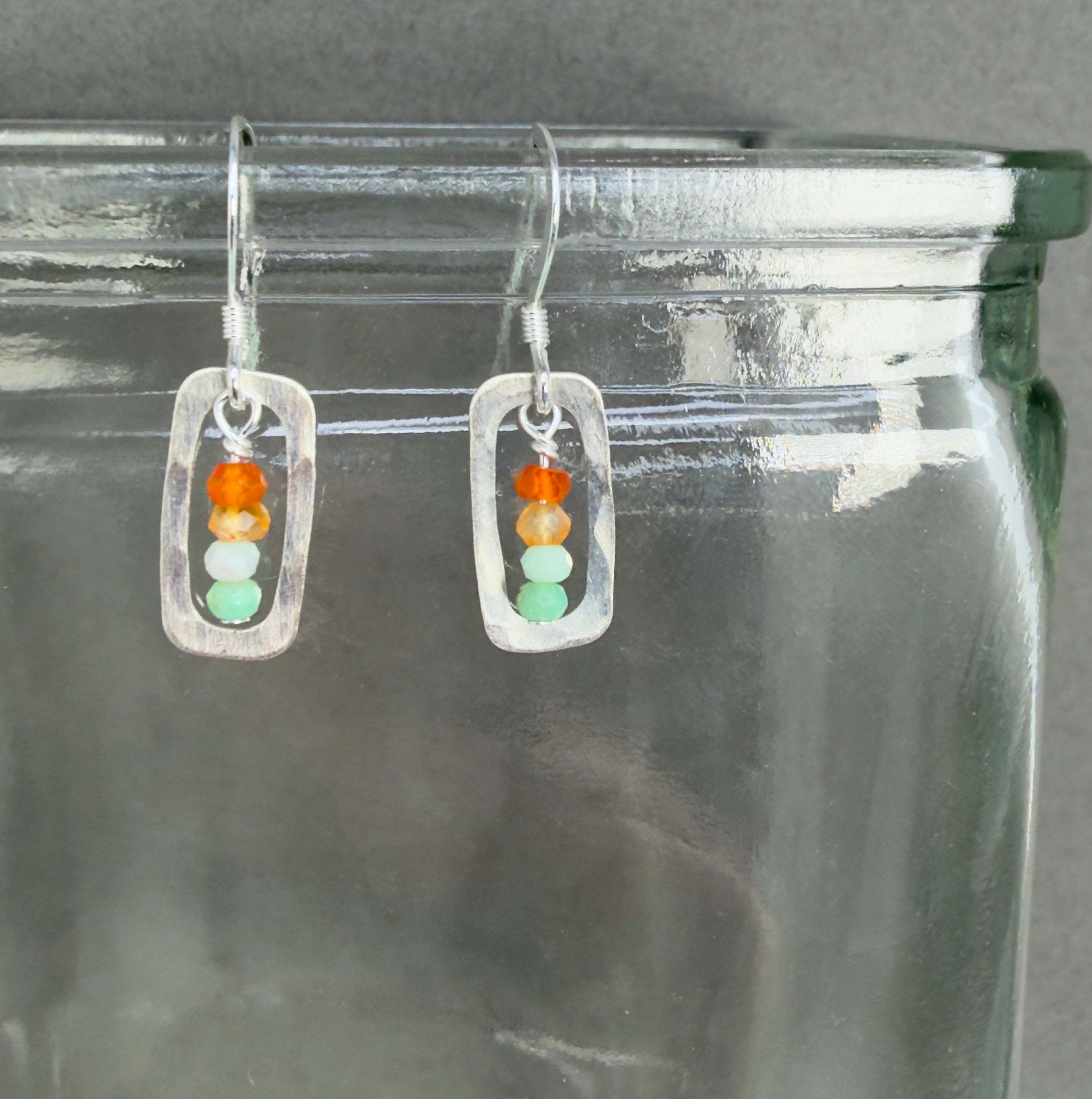 Pair of earrings with a stack or orange, pale orange, pale minty green and medium minty green beads framed with a rounded silver rectangular frame, on sterling silver hooks. They're shown hanging on a clear glass jar with that hint of green clear glass has, in front of a dark grey background.