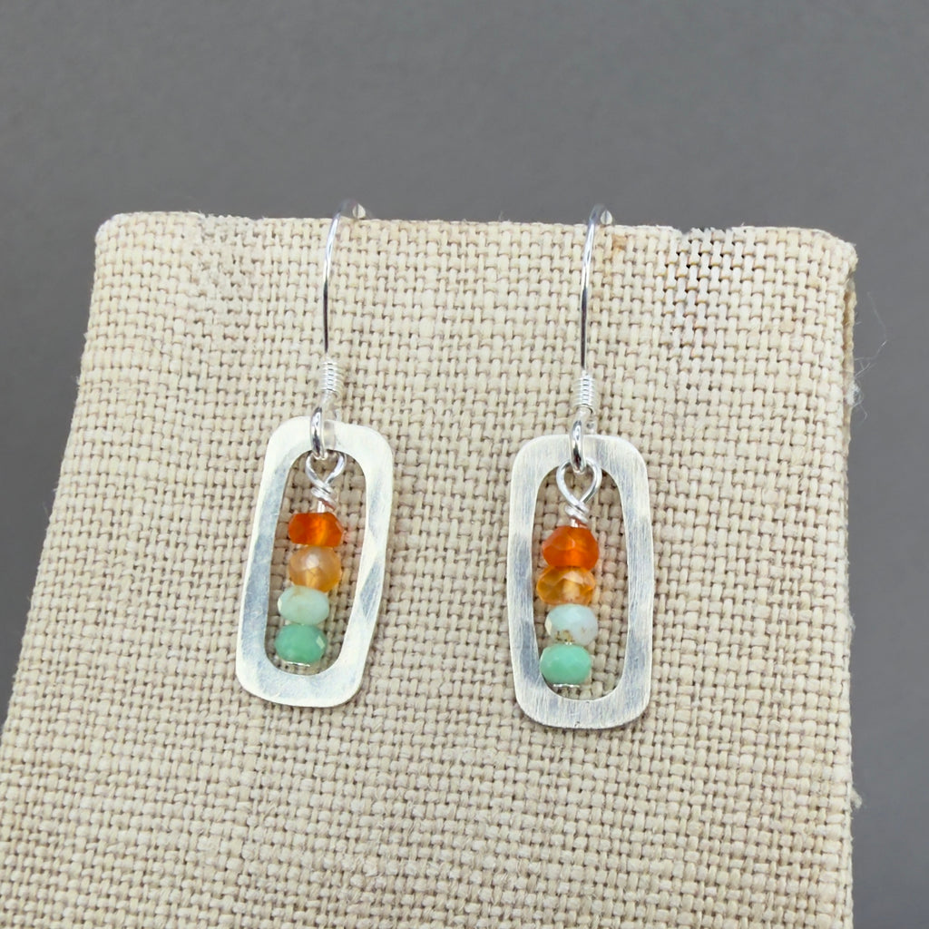 Pair of earrings with a stack or orange, pale orange, pale minty green and medium minty green beads framed with a rounded silver rectangular frame, on sterling silver hooks. Shown on a beige linen display which really makes the colors pop. These earrings hang 1 1/8" inch or about 3cm from the piercing.