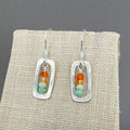 Pair of earrings with a stack or orange, pale orange, pale minty green and medium minty green beads framed with a rounded silver rectangular frame, on sterling silver hooks. Shown on a beige linen display which really makes the colors pop. These earrings hang 1 1/8" inch or about 3cm from the piercing.