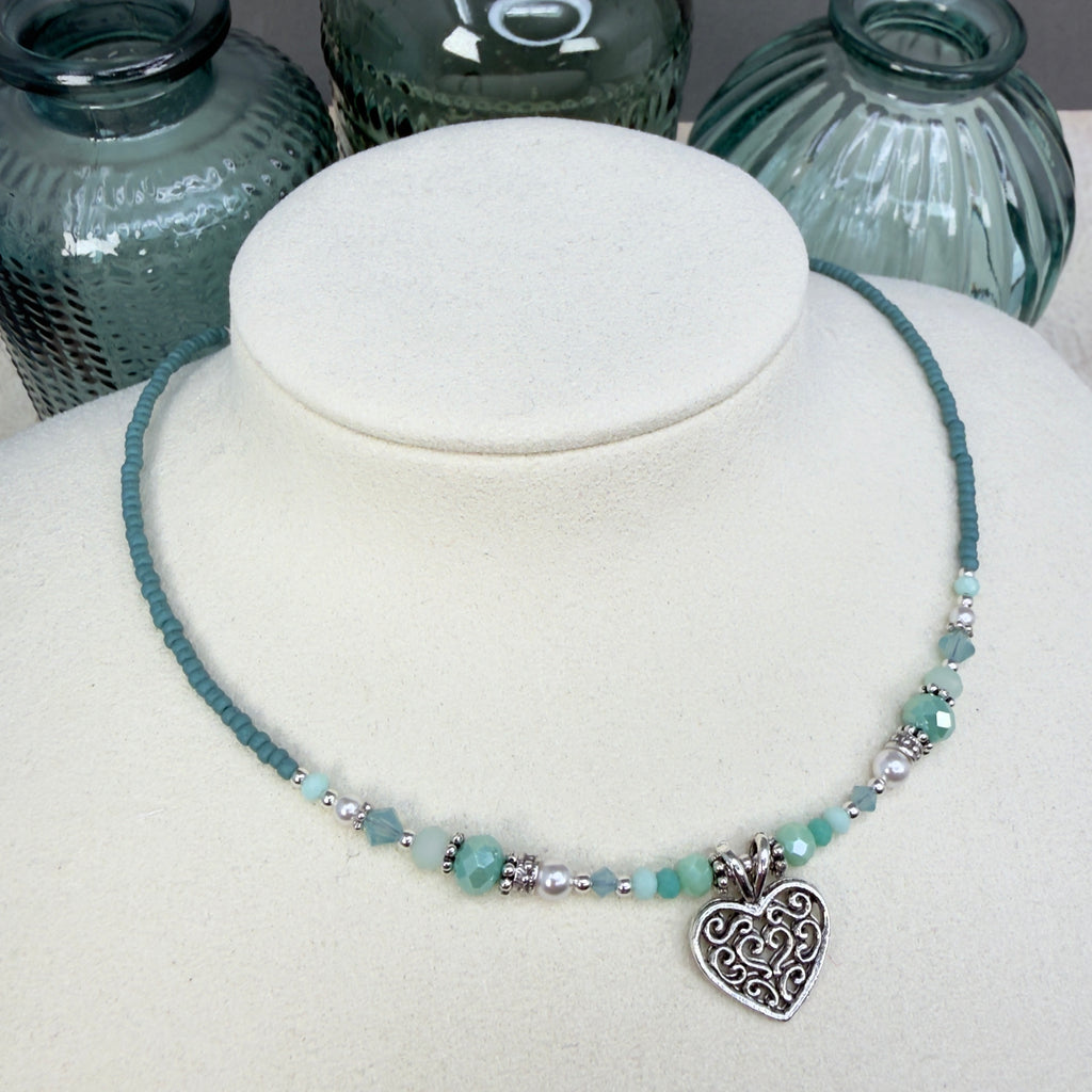 Soft minty green and white with silver accents and a heart pendant on an ivory mannequin bust against a neutral background with green glass bottles