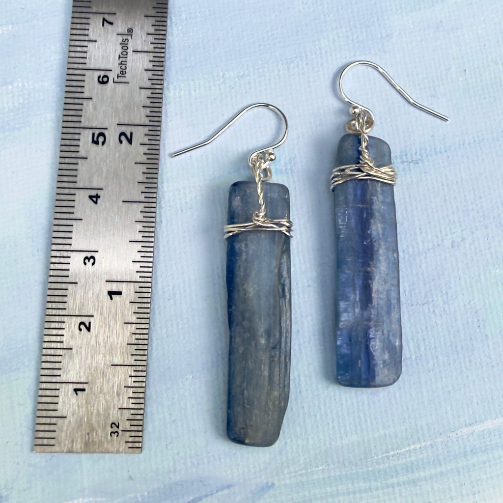 A pair of handmade dangle earrings made from natural blue-gray kyanite chips wrapped with silver wire, shown next to a metal ruler on a light blue textured background. Earrings measure 2 1/4" length or 56mm.