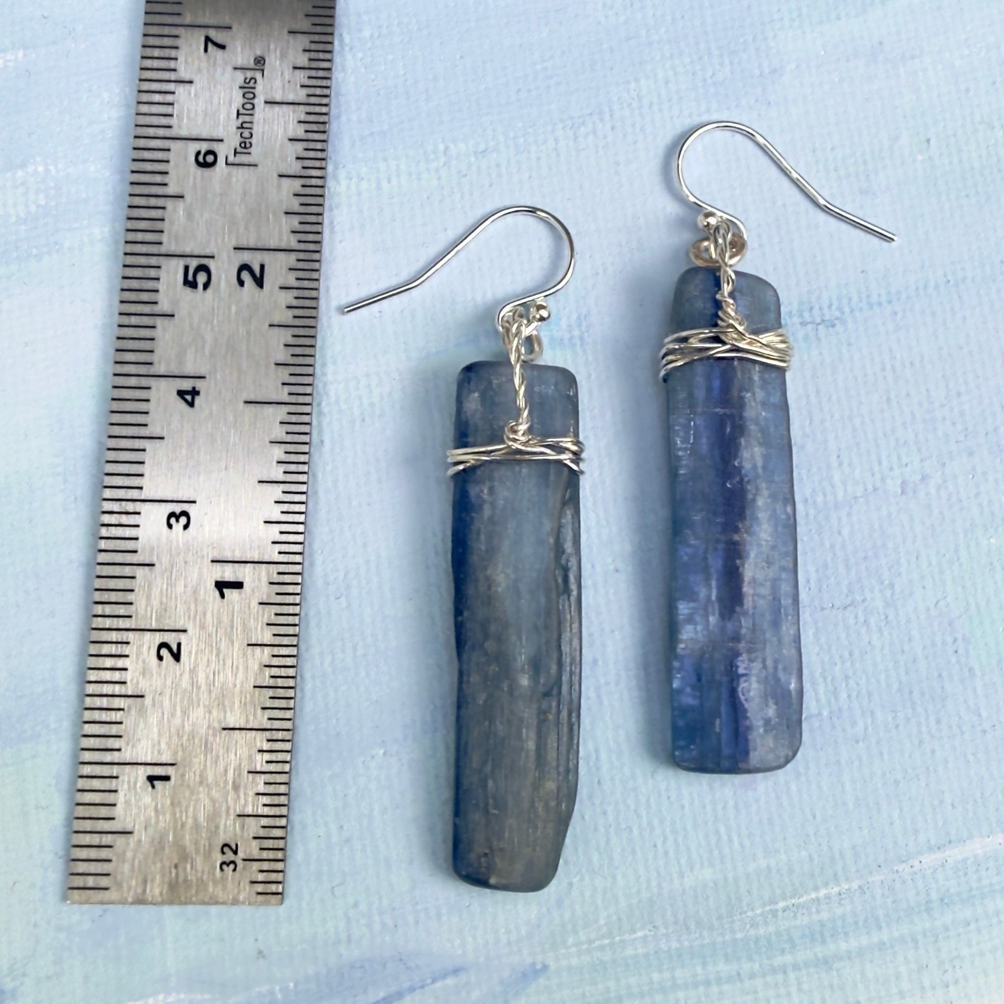 A pair of handmade dangle earrings made from natural blue-gray kyanite chips wrapped with silver wire, shown next to a metal ruler on a light blue textured background. Earrings measure 2 1/4" length or 56mm.