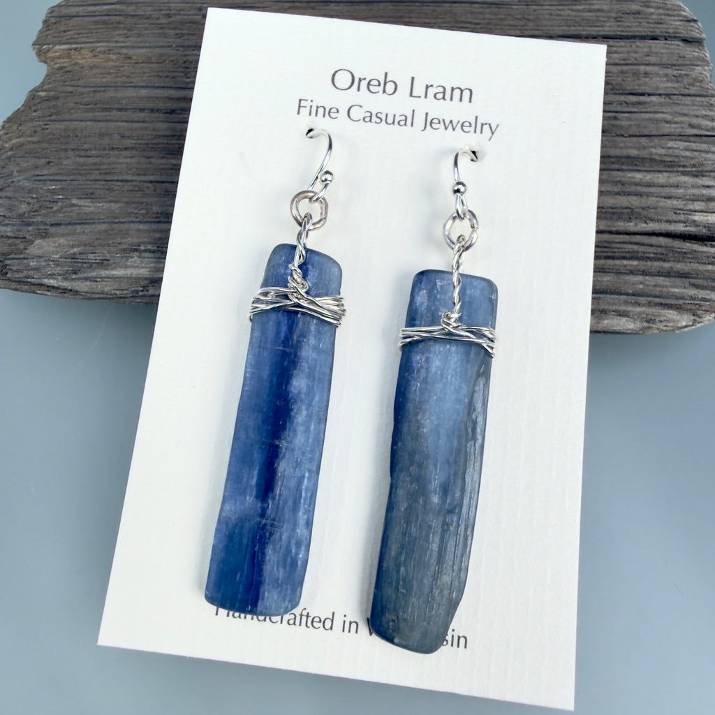A pair of blue stone dangle earrings with silver wire wrapping and silver fishhook ear wires, displayed on a white jewelry card labeled 'Oreb Lram Fine Casual Jewelry' against a rustic wood background.
