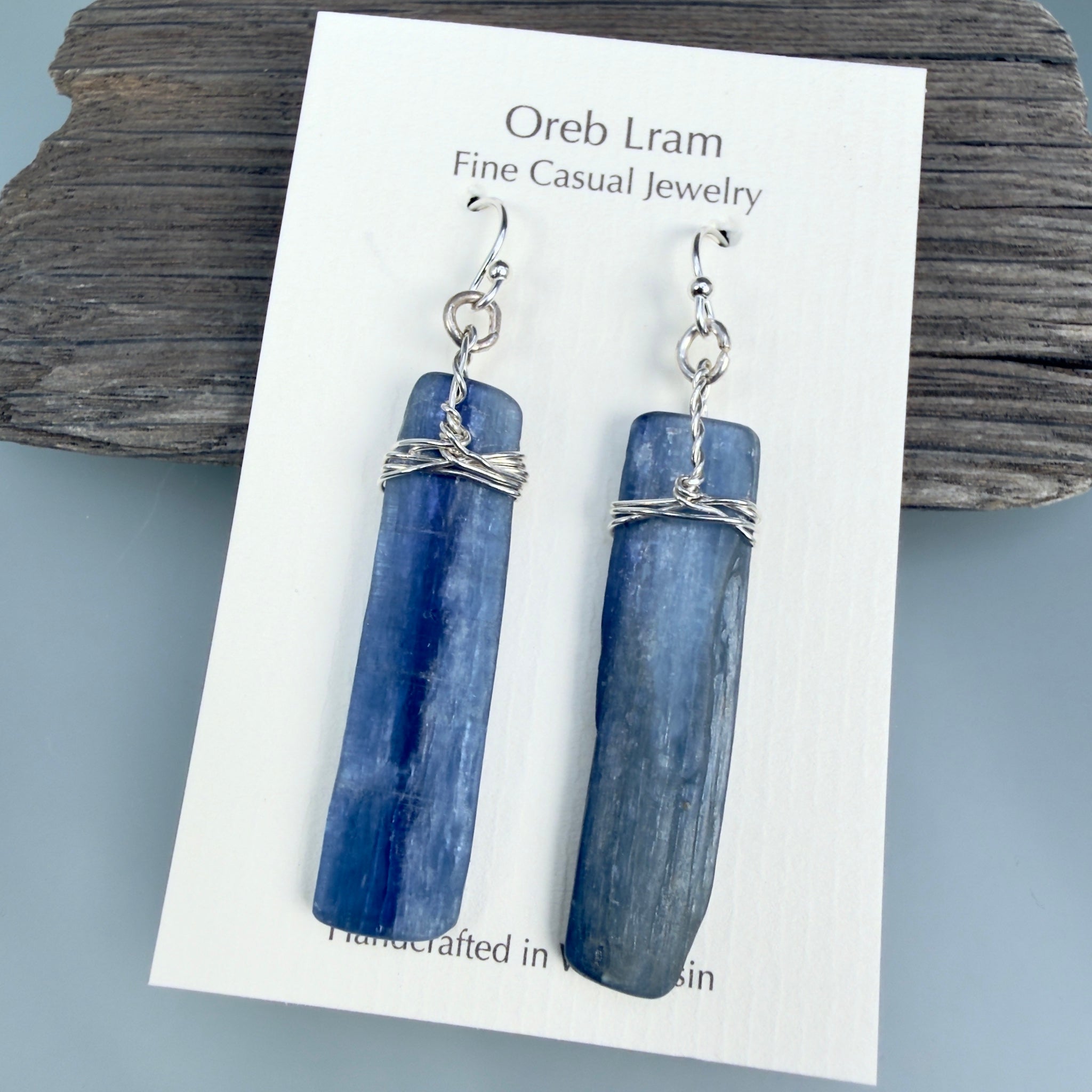 A pair of blue stone dangle earrings with silver wire wrapping and silver fishhook ear wires, displayed on a white jewelry card labeled 'Oreb Lram Fine Casual Jewelry' against a rustic wood background.