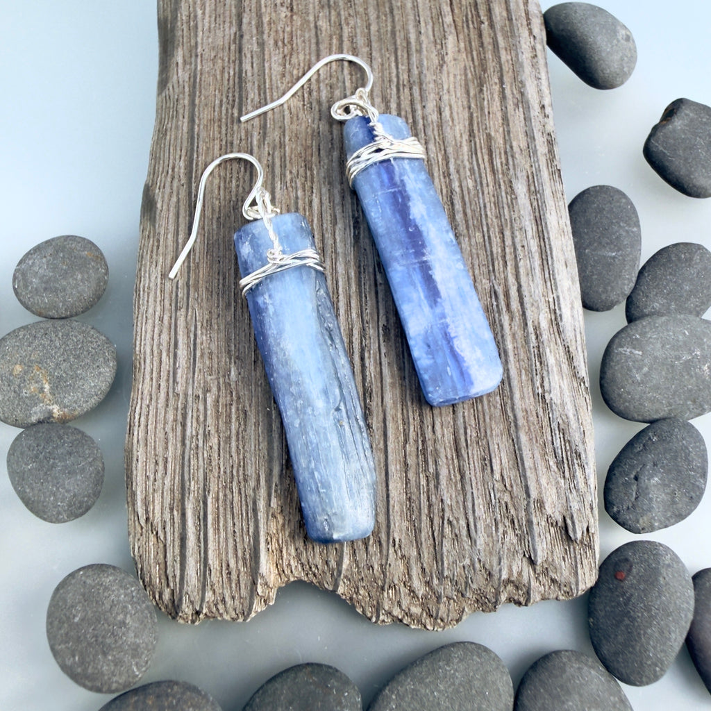 Flat blue grey Kyanite gemstones wrapped in silver at the top and hanging from sterling silver hooks. The stones are rough cut rectangular slabs with minimal polish. Displayed on a dark grey piece of driftwood and surrounded with grey pebbles on a light grey background.