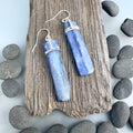 Flat blue grey Kyanite gemstones wrapped in silver at the top and hanging from sterling silver hooks. The stones are rough cut rectangular slabs with minimal polish. Displayed on a dark grey piece of driftwood and surrounded with grey pebbles on a light grey background.