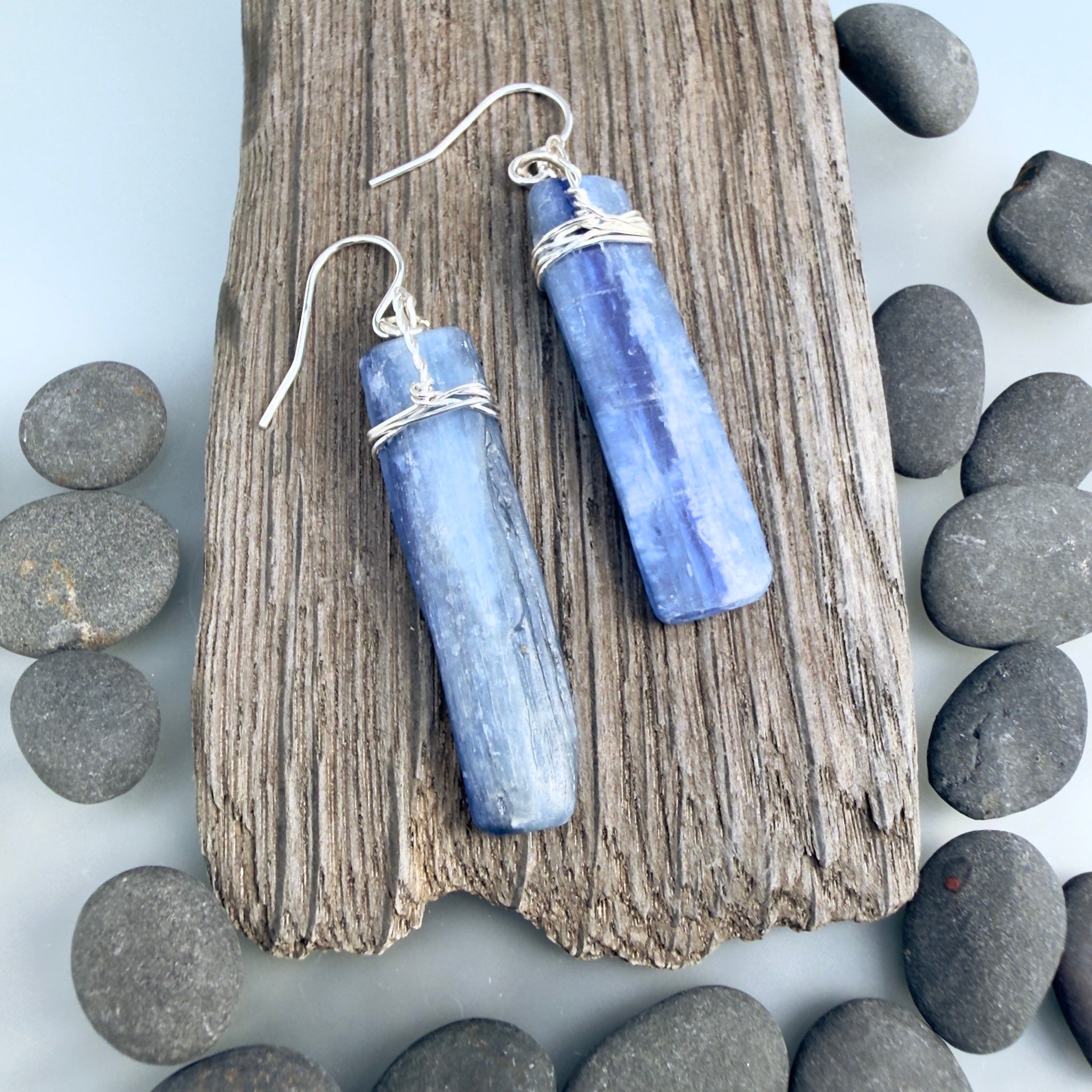 Flat blue grey Kyanite gemstones wrapped in silver at the top and hanging from sterling silver hooks. The stones are rough cut rectangular slabs with minimal polish. Displayed on a dark grey piece of driftwood and surrounded with grey pebbles on a light grey background.
