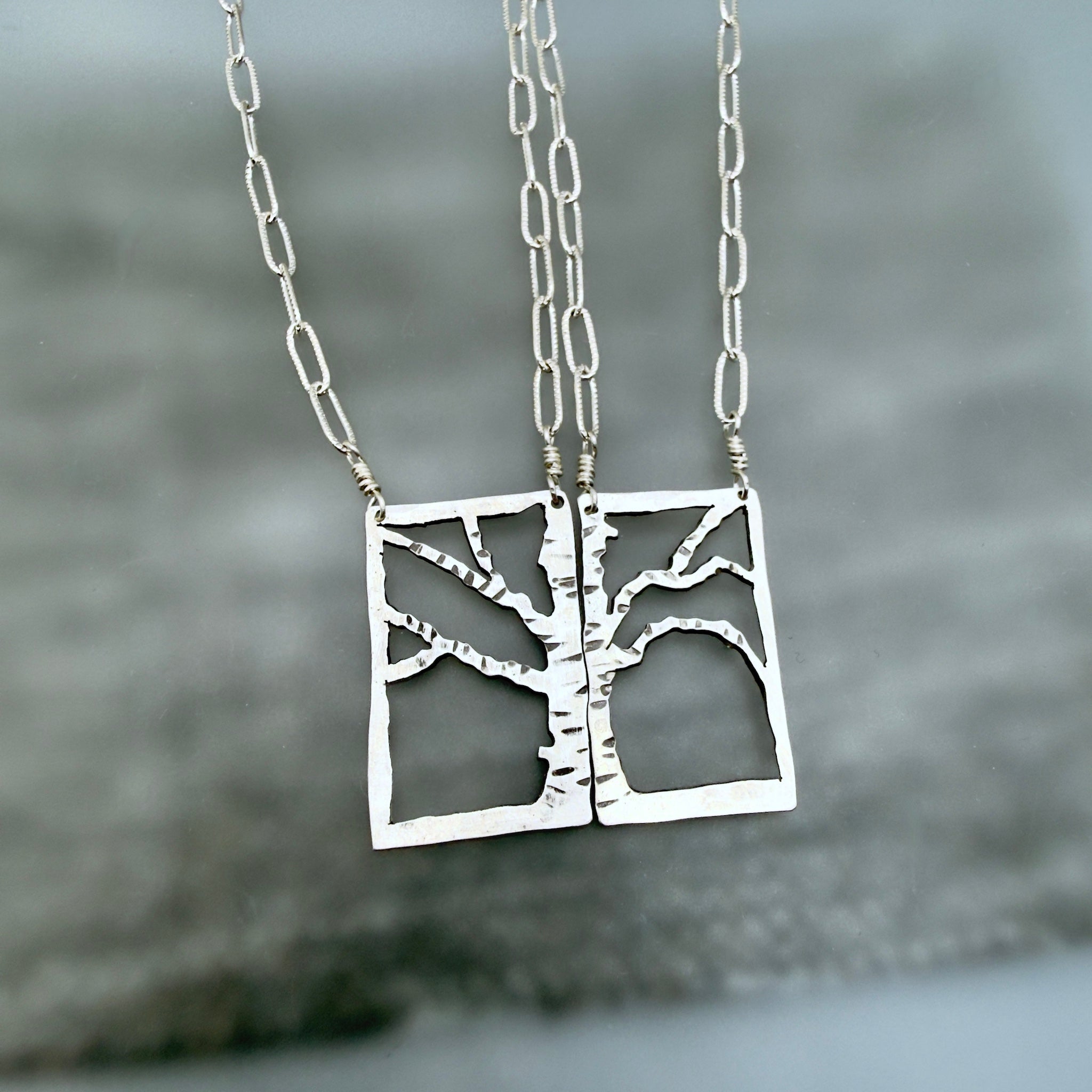 A pair of silver necklaces with square pendant cutouts featuring a tree design, hanging against a blurred gray background.