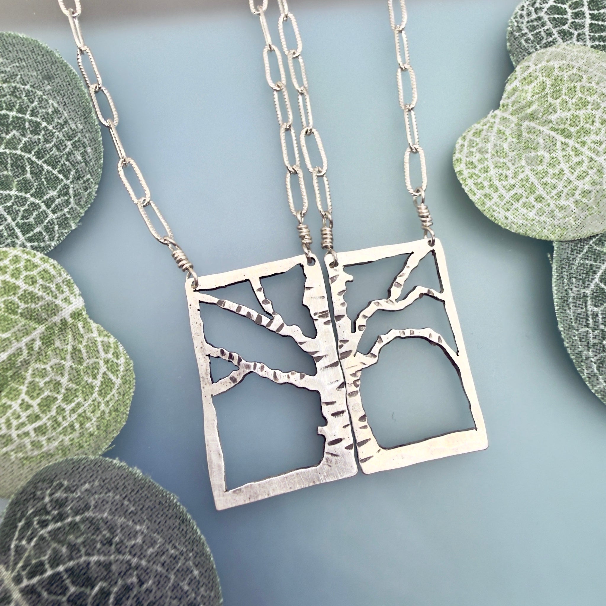 Two matching silver tree-of-life pendant necklaces with paperclip chains, displayed on a light blue background with decorative green leaves.