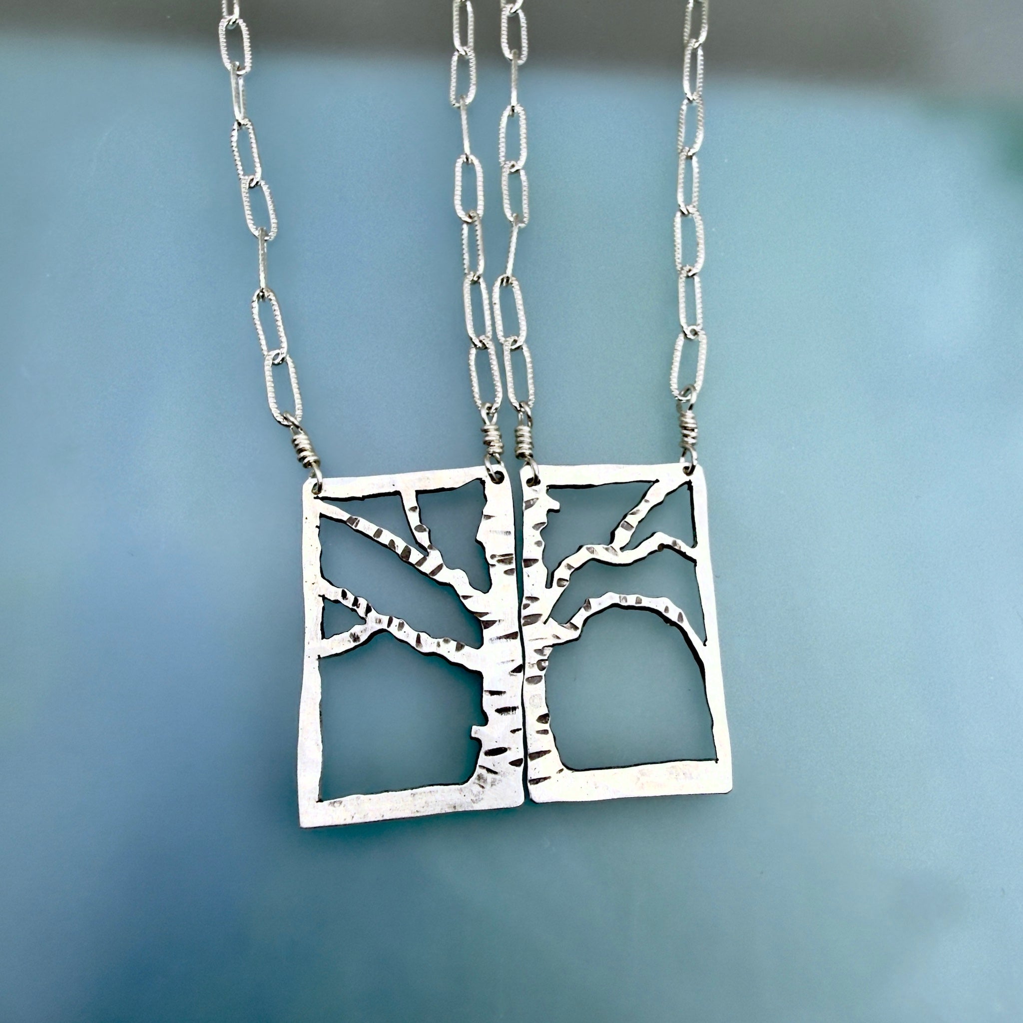 Two silver tree-of-life pendant necklaces with rectangular cutout pendants and chain links, displayed against a soft blue background.