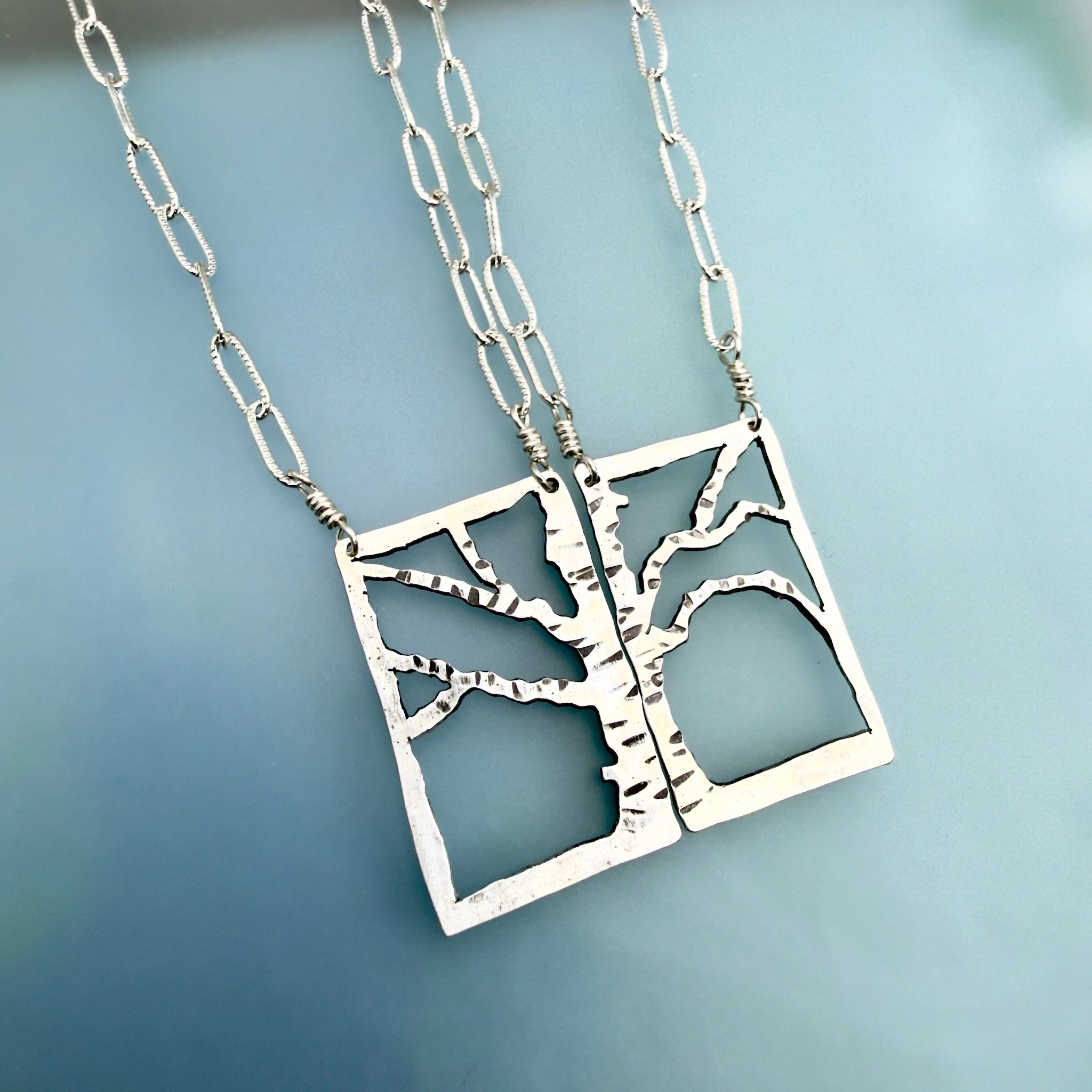 Two silver tree-of-life pendant necklaces with square cutout pendants and chain links, displayed on a soft blue background.