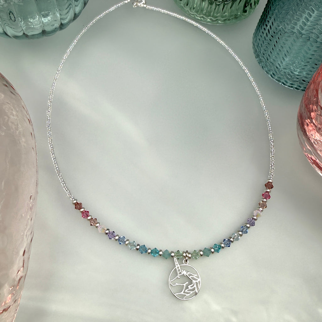 Necklace with multicolored beads and a silver unicorn charm on a light gray surface with pastel colored glass bottles at the edges of the picture.