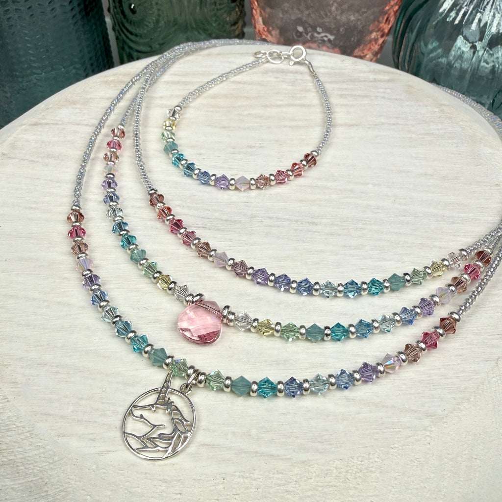 3 Multicolored pastel beaded necklaces layered, and a matching bracelet, on a textured white surface. This picture is showing the various items available in this line. The front necklace is the unicorn version, the middle necklace is the same style but with a pink crystal drop at center instead of the unicorn, and the back necklace has no pendant drop, only in line pastel crystals. The bracelet is the same style as the back necklace but matches all of them. 