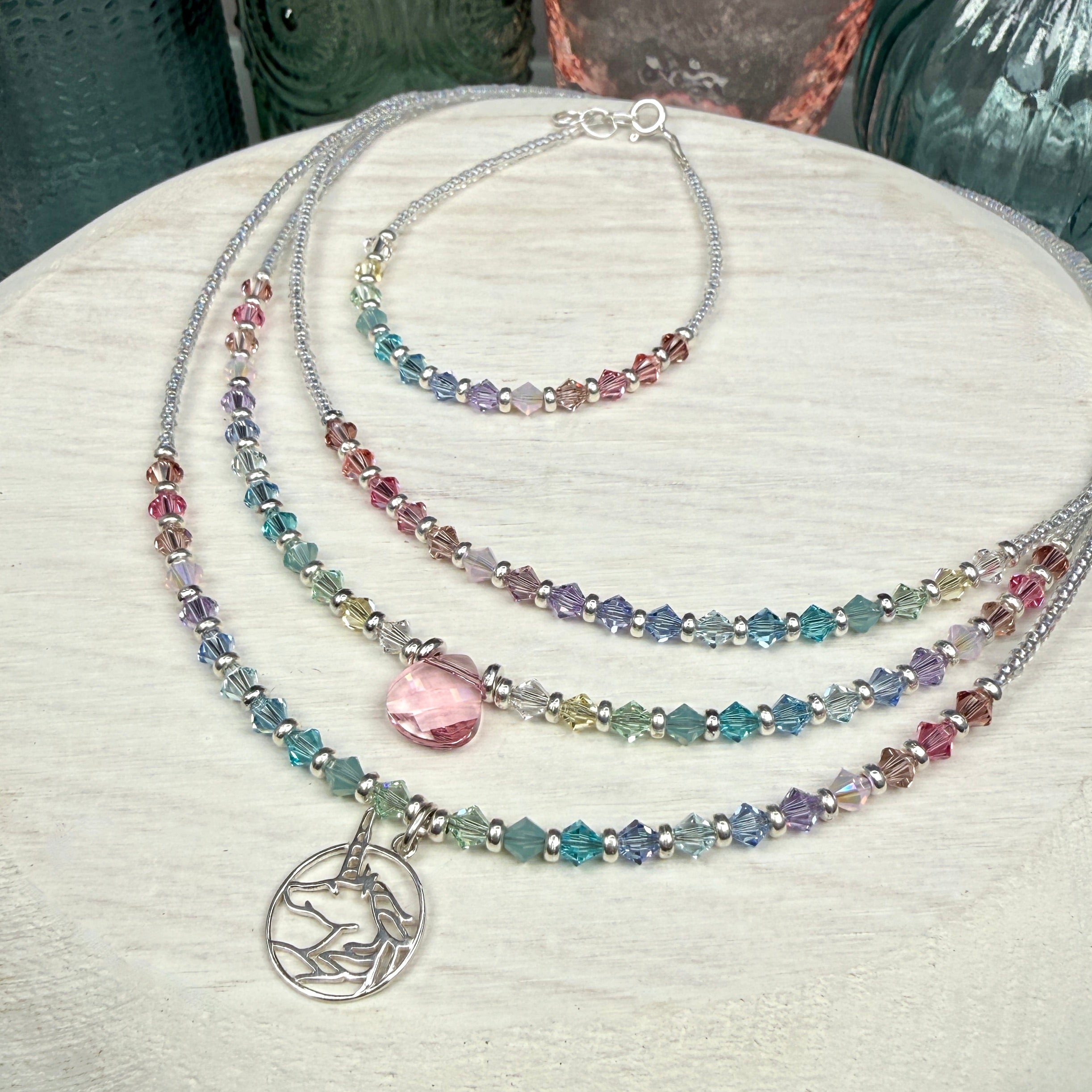 3 Multicolored pastel beaded necklaces layered, and a matching bracelet, on a textured white surface. This picture is showing the various items available in this line. The front necklace is the unicorn version, the middle necklace is the same style but with a pink crystal drop at center instead of the unicorn, and the back necklace has no pendant drop, only in line pastel crystals. The bracelet is the same style as the back necklace but matches all of them. 