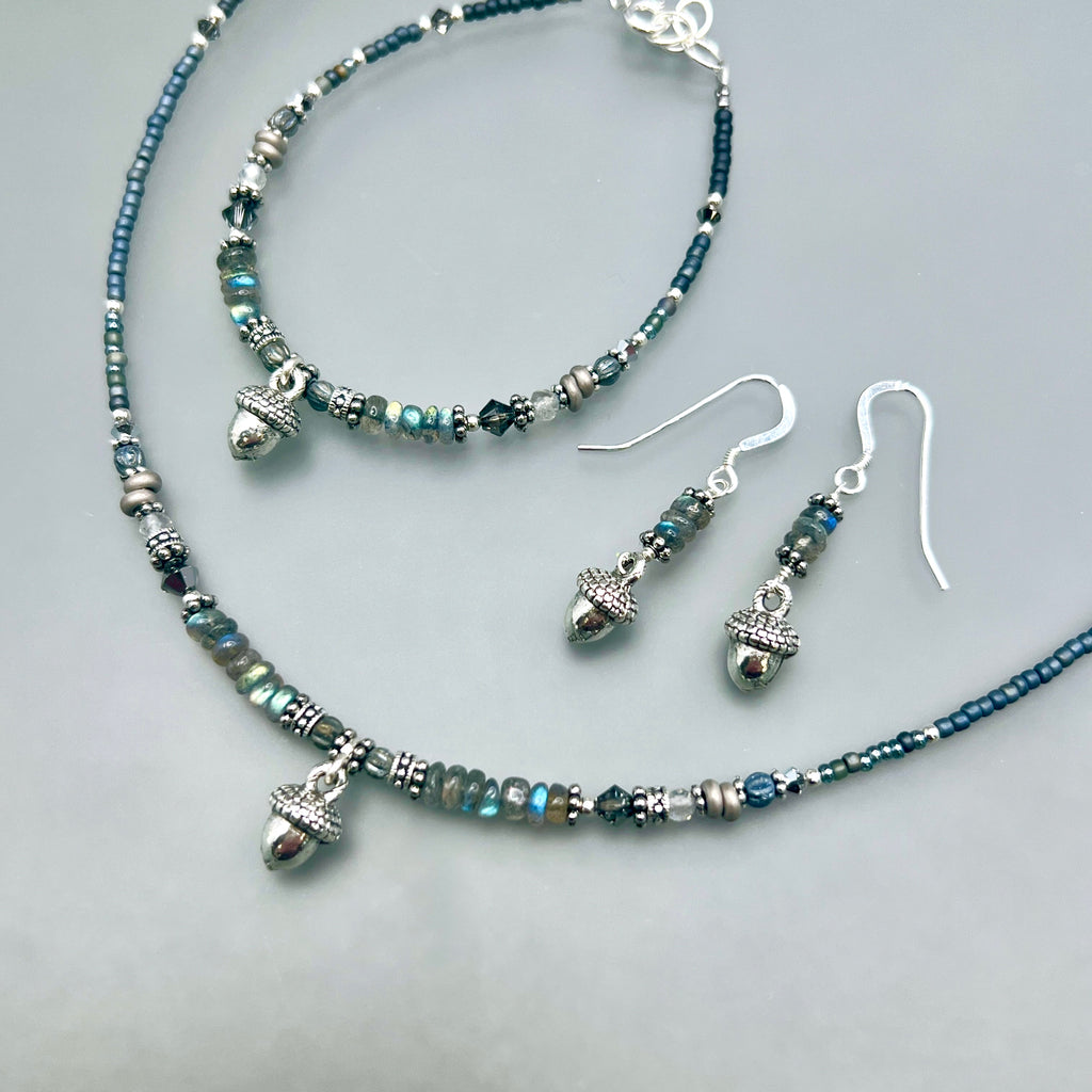 Grey and silver Jewelry set with necklace, bracelet, and earrings on a light grey background. Each piece has a small silver acorn and natural iridescent labradorite gemstones.