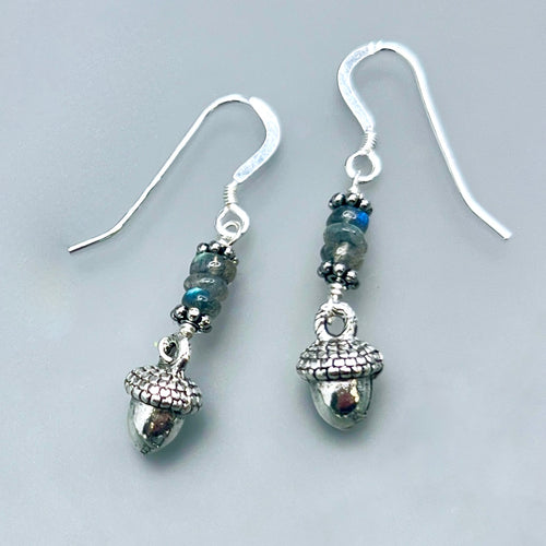 Grey and silver earrings on a pale grey background. The earrings feature a stack of natural grey labradorite gemstone with flashes of blue iridescence, with small silver pewter acorns beneath.