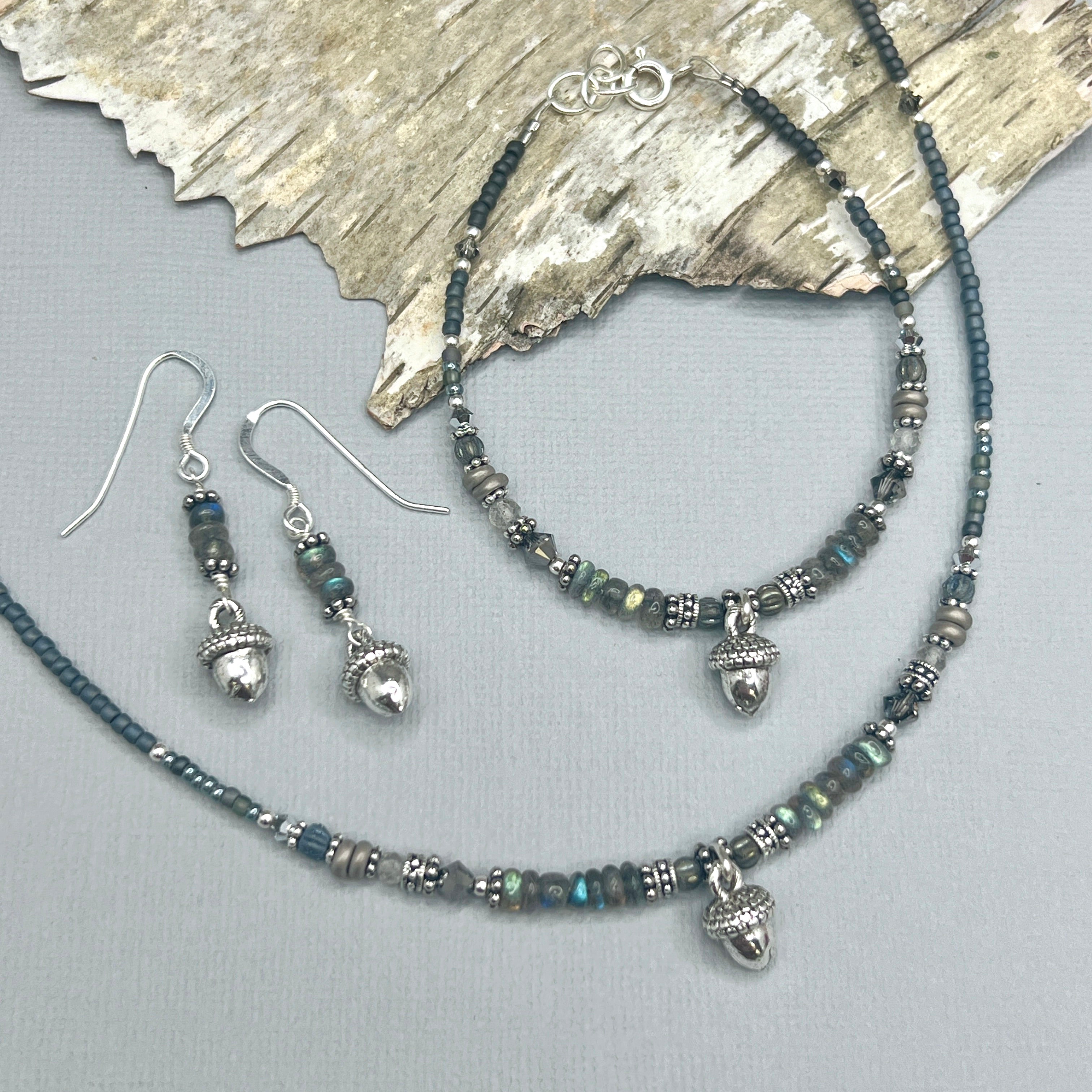 Grey and silver Jewelry set with necklace, bracelet, and earrings on a textured surface. Each piece has a small silver acorn and natural iridescent labradorite gemstones.